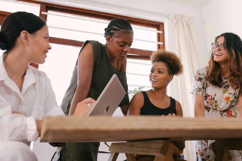 A group of women sitting around a table looking at a laptop talking about IT consulting services with a client.